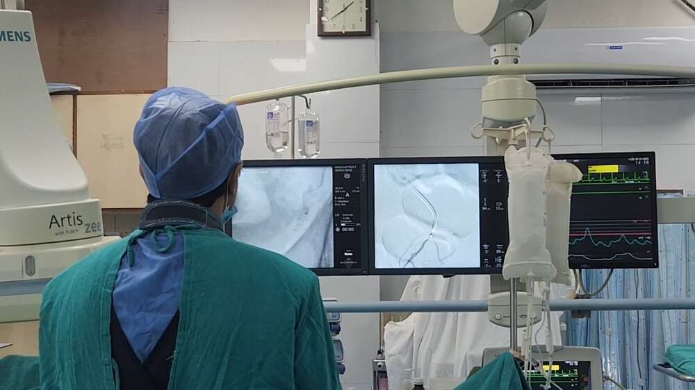 Doctor performing prostate artery embolization procedure, viewing real-time angiography images on monitors in an interventional radiology lab